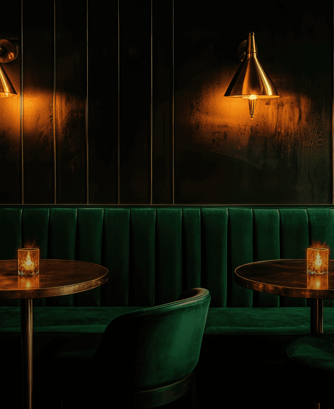 Intimate bar seating with emerald green velvet banquette, brass cocktail tables, candlelight, and warm wall sconces creating a refined hospitality atmosphere.